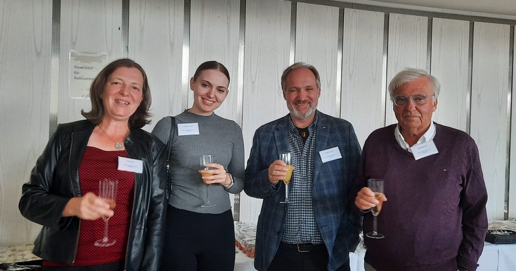 Vier Personen bei einer Veranstaltung stoßen gemeinsam mit Getränken an und lächeln in die Kamera.